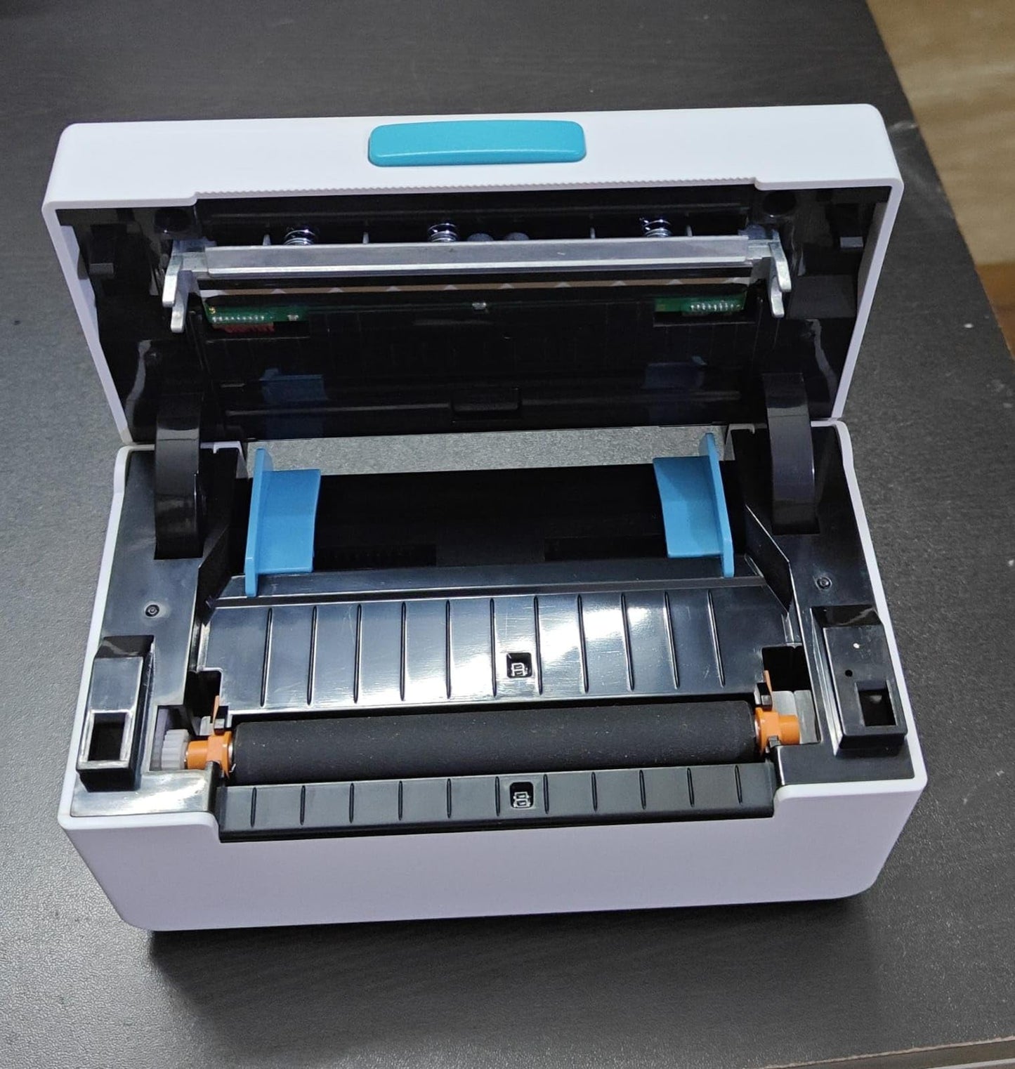 Open the KUPIS KP410 4x6 Shipping Label Printer on a dark surface to highlight its quality printing. Inside, view the easy-to-load rollers and label slot. The white exterior is accented by a teal button on top, ideal for eCommerce barcode use.