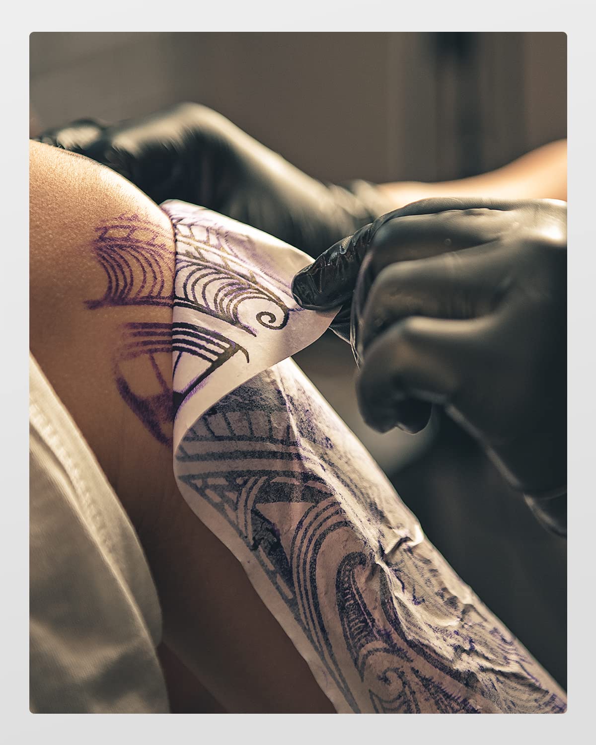 A tattoo artist in black gloves carefully removes the Phomemo A4 Tattoo Transfer Paper, compatible with M08F, from a clients arm to reveal a newly inked intricate design of swirls and patterns.