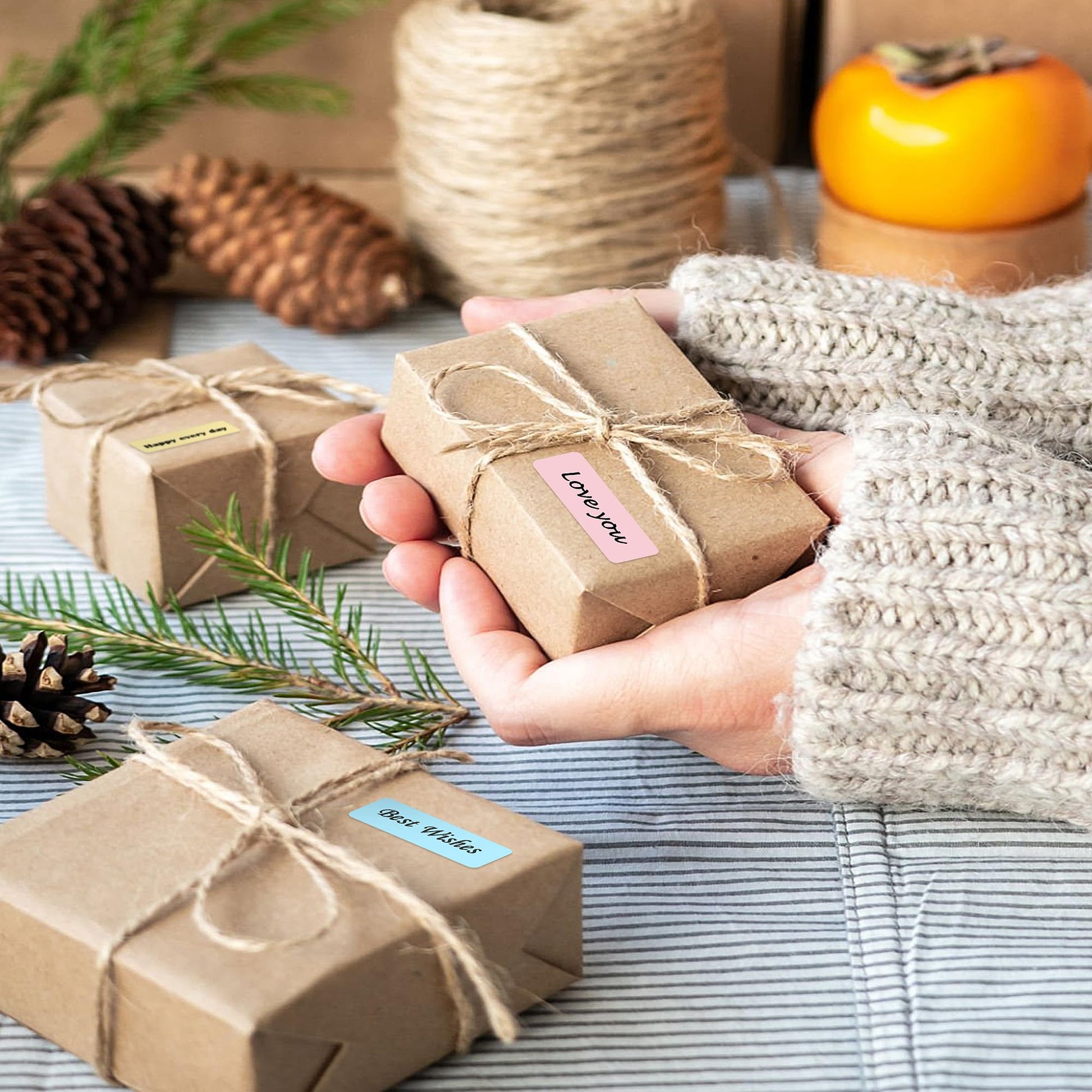 A person in a cozy sweater holds a small gift wrapped in brown paper with twine, labeled Love You using the POLONO P31S Label Maker. Nearby are similar gifts, pinecones, twine, and a persimmon on a striped cloth surface.